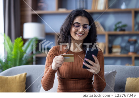 Close-up photo of a young smiling Muslim girl at home, sitting on the sofa, holding a credit card and using a mobile phone. 122767349