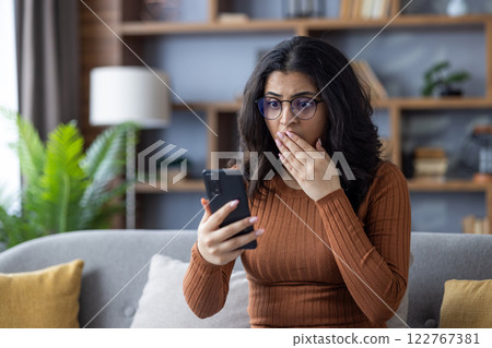 Close-up photo of a sad young Indian woman sitting on the sofa at home, looking at the phone in shock, covering her mouth with her hand. Close-up photo of a sad young Indian woman sitting on the sofa at home, looking at the phone in shock, covering her mouth with her hand. 122767381