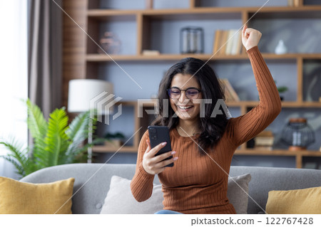 Happy young Indian girl sitting on sofa at home, looking at mobile phone screen, reading good news, raised hand up celebrating. Happy young Indian girl sitting on sofa at home, looking at mobile phone screen, reading good news, raised hand up celebrating. 122767428