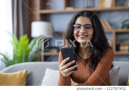Close-up portrait of young Muslim woman sitting on sofa at home, holding phone in hand and looking at camera. 122767432