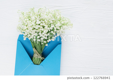 delicate bouquet of lily of the valley flowers tied with a ribbon, placed inside a bright blue envelope on a white wooden background, symbolizing love and appreciation 122768941
