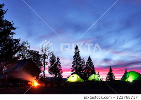 Night camping. Tourist have a rest at a campfire near illuminated tent and wooden house under amazing night sky full of stars and milky way Night camping. Tourist have a rest at a campfire near illuminated tent and wooden house under amazing night sky full of stars and milky way 122769717