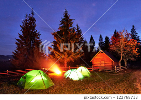 Night camping. Tourist have a rest at a campfire near illuminated tent and wooden house under amazing night sky full of stars and milky way. Astrophotography Night camping. Tourist have a rest at a campfire near illuminated tent and wooden house under amazing night sky full of stars and milky way. Astrophotography 122769718