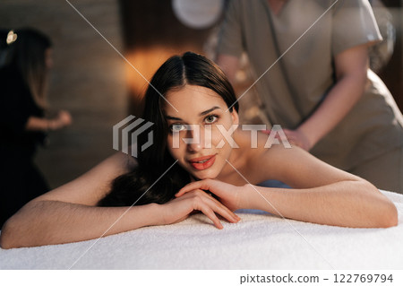 Portrait of pretty woman relaxing during back massage in spa with soft light, looking at camera. Beautiful naked female with perfect skin getting relaxing massage. Concept of rest with spa treatment. Portrait of pretty woman relaxing during back massage in spa with soft light, looking at camera. Beautiful naked female with perfect skin getting relaxing massage. Concept of rest with spa treatment. 122769794