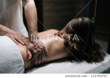 Close-up hands of masseur male performing deep tissue technique on female lower back, targeting muscle tension and promoting wellness during bodywork session in beauty parlor with dark interior. Close-up hands of masseur male performing deep tissue technique on female lower back, targeting muscle tension and promoting wellness during bodywork session in beauty parlor with dark interior. 122769797