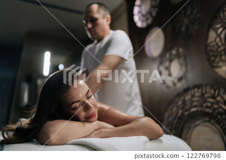 Calm happy young woman relaxing during back massage in spa salon, lying with closed eyes and smiling, close-up. Massage therapist providing therapeutic back massage to serene client, clsoeup. 122769798