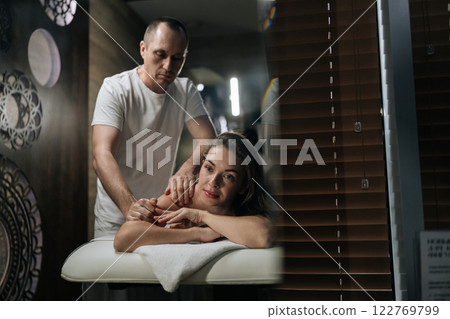 Portrait of beautiful young woman relaxing during back massage in spa with soft light, looking at camera. Charming brunette female getting relaxing massage. Concept of rest with spa treatment. 122769799