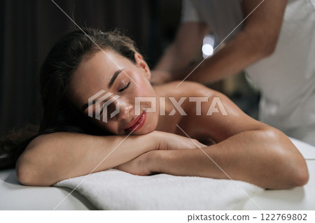 Close-up face of happy relaxed woman getting luxury back massage lying on massage table closed eyes. Professional masseur performing therapeutic massage on relaxed female client. 122769802
