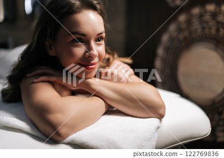 Close-up face of smiling young woman lying on massage table with hands under chin preparing for relax massage procedure in spa salon with dark interior. Concept of spa health and self-care. Close-up face of smiling young woman lying on massage table with hands under chin preparing for relax massage procedure in spa salon with dark interior. Concept of spa health and self-care. 122769836