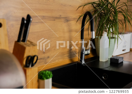 Side view of the kitchen black granite sink on with flexible black faucet on the background of wooden countertop and kitchen utensils and plants. Kitchen island, interior, dishwashing concept. High Side view of the kitchen black granite sink on with flexible black faucet on the background of wooden countertop and kitchen utensils and plants. Kitchen island, interior, dishwashing concept. High 122769935