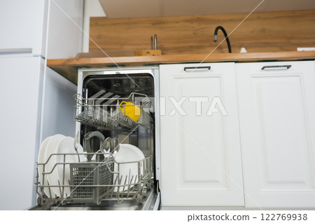 An open dishwasher with a dish drawer pulled out close-up on the left, it contains clean dishes and cutlery against the background of a white wooden kitchen. Comfort concept, dishwashing. High quality An open dishwasher with a dish drawer pulled out close-up on the left, it contains clean dishes and cutlery against the background of a white wooden kitchen. Comfort concept, dishwashing. High quality 122769938