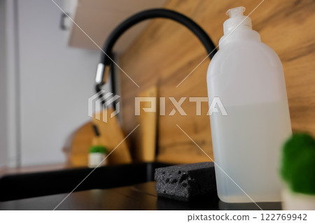 Close-up of a white bottle of dishwashing soap, a black dishwashing sponge lying next to it and a potted plant against a background of a black granite sink and a flexible high faucet and a white 122769942