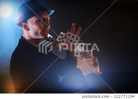 Magician showing trick with playing cards. Magic or dexterity, circus, gambling. Prestidigitator in dark room with fog Magician showing trick with playing cards. Magic or dexterity, circus, gambling. Prestidigitator in dark room with fog 122771194