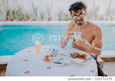 Breakfast near the swimming pool. Young man on vacation in hotel having breakfast near swimming pool. Person having breakfast in the hotel with pool in the background Breakfast near the swimming pool. Young man on vacation in hotel having breakfast near swimming pool. Person having breakfast in the hotel with pool in the background 122772570
