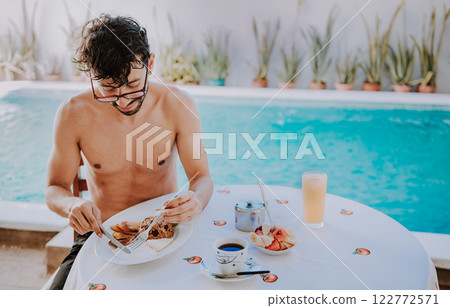 Young man on vacation in hotel having breakfast near swimming pool. Breakfast near the swimming pool. Person having breakfast in the hotel with pool in the background Young man on vacation in hotel having breakfast near swimming pool. Breakfast near the swimming pool. Person having breakfast in the hotel with pool in the background 122772571