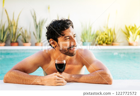 Portrait of man at the edge of swimming pool with a glass of wine. Handsome man in swimming pool holding glass of wine. Handsome guy on the edge of the pool holding a glass of wine 122772572