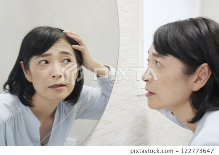 一位 50 多歲的女性看著鏡子，擔心白髮。 122773647
