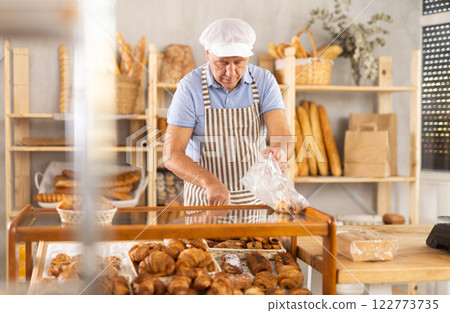 Senior man seller puts croissant in plastic disposable bag for buyer, packs goods. 122773735