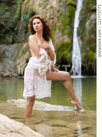 Half-naked girl in white translucent negligee poses standing in nature against backdrop of waterfall 122773773