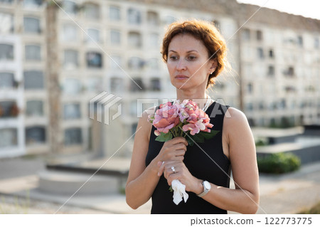 Woman in black dress with bouquet of flowers came to cemetery to honor the memory of deceased loved one 122773775