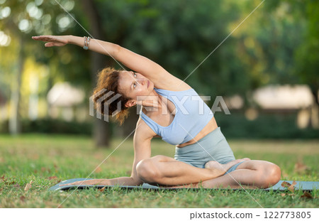 Woman practicing hatha yoga in garden, stretching in an asymmetrical seated asana Parivritta Janu Sirsasana Woman practicing hatha yoga in garden, stretching in an asymmetrical seated asana Parivritta Janu Sirsasana 122773805