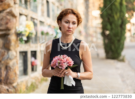 Sad woman in black dress with bouquet of flowers and handkerchief in hands, stands in cemetery Sad woman in black dress with bouquet of flowers and handkerchief in hands, stands in cemetery 122773830
