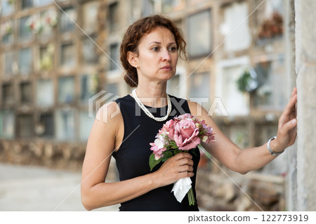 Grieving woman in black standing with bouquet near columbarium 122773919