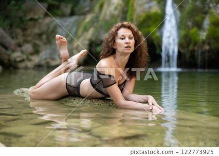 Woman in mesh black bikini sits on rock on shore of tropical waterfall Woman in mesh black bikini sits on rock on shore of tropical waterfall 122773925