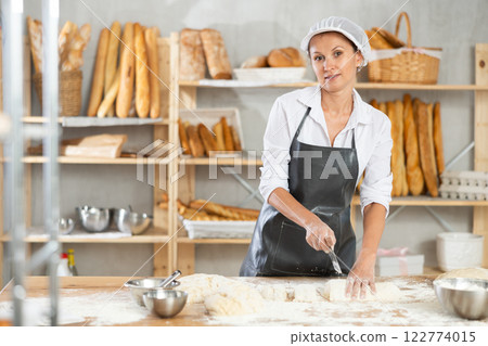 Woman works in bakery as baker, cuts dough into portions, forms pieces of dough to create buns. Woman works in bakery as baker, cuts dough into portions, forms pieces of dough to create buns. 122774015