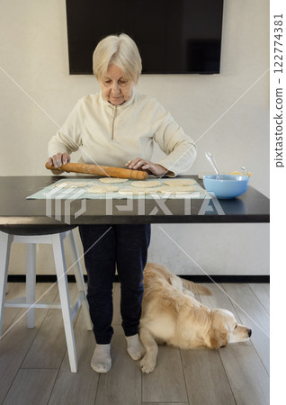 An elderly woman rolls out dough on a kitchen counter while her Golden Retriever lies on the floor beside her, resting peacefully in a cozy home environment. An elderly woman rolls out dough on a kitchen counter while her Golden Retriever lies on the floor beside her, resting peacefully in a cozy home environment. 122774381