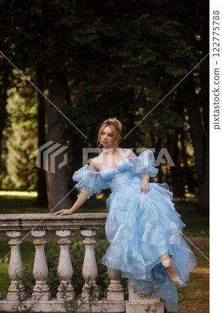 Romantic Woman in a Light Blue Gown Posing on a stone balustrade in a picturesque garden. Romantic Woman in a Light Blue Gown Posing on a stone balustrade in a picturesque garden. 122775788