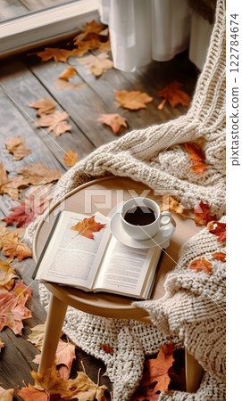 Cozy autumn setting with fall leaves, a steaming cup of coffee, an open book, and a knitted blanket. Inviting comfort concept 122784674
