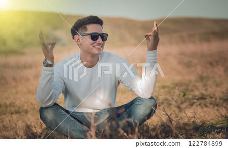 Smiling young man sitting on the grass, smiling young man in eyeglasses sitting on the field, concept of happiness in the field 122784899