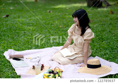 Young woman is sitting on a cloth on the grass with food and snacks Young woman is sitting on a cloth on the grass with food and snacks 122785637