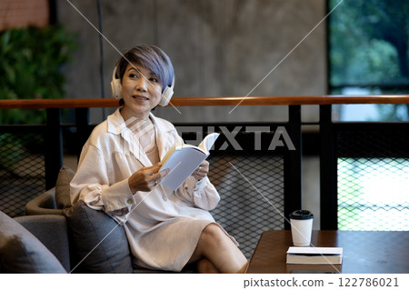 Elder woman sitting on sofa  wearing headphones reading a book 122786021