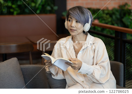 Elder woman sitting on sofa  wearing headphones reading a book 122786025