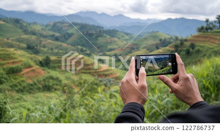 Hands holding smartphone capturing stunning mountain landscape with terraced rice fields. Travel photography of scenic Asian agricultural vista with rolling hills and cloudy sky 122786737