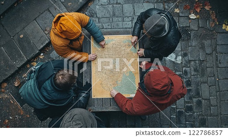 Aerial view of four people studying vintage map together on dark cobblestone ground. Friends in colorful winter jackets planning route during autumn outdoor adventure 122786857