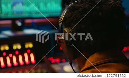 African american tracking engineer processing and mixing sounds in control room, operating audio console with moving faders and colored meters. Technician producer deals with technical gear. Camera A. 122791832