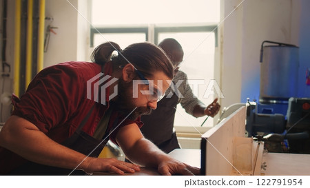 Woodworking expert wears protection gear, inserting plank in spindle moulder next to coworker. CNC machinist uses safety glasses while cutting lumber with wood shaper helped by colleague, camera B Woodworking expert wears protection gear, inserting plank in spindle moulder next to coworker. CNC machinist uses safety glasses while cutting lumber with wood shaper helped by colleague, camera B 122791954