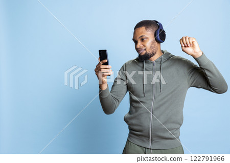 Carefree relaxed man listening to funky cool music from his playlist, using smartphone and headphones to dance and enjoy their free time. Euphoric person raving on rhythm, doing dance movements. 122791966
