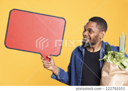 African american guy holds a speech bubble empty icon to create an ad for zero waste and sustainable lifestyle. Doing a promotional marketing advertisement for fresh bio fruits and veggies. African american guy holds a speech bubble empty icon to create an ad for zero waste and sustainable lifestyle. Doing a promotional marketing advertisement for fresh bio fruits and veggies. 122792031