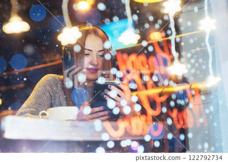 Young beautiful woman sitting in cafe, drinking coffee. Model listening to music. Christmas, Happy new year, Valentines day, winter holidays concept Young beautiful woman sitting in cafe, drinking coffee. Model listening to music. Christmas, Happy new year, Valentines day, winter holidays concept 122792734