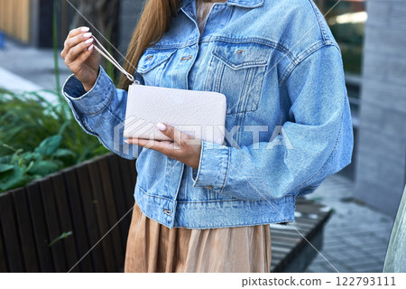 Woman wearing denim jacket holding white wallet outdoors showing casual style and fashion accessory concept Woman wearing denim jacket holding white wallet outdoors showing casual style and fashion accessory concept 122793111