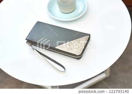 Elegant leather wallet with intricate metal detail on white table, next to cup and saucer, fashion accessory style concept 122793140