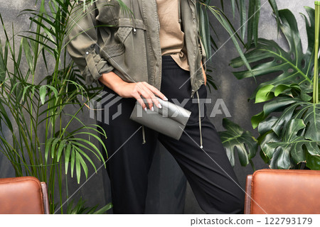 Elegant woman in khaki jacket holding stylish clutch amidst lush green plants, modern fashion concept Elegant woman in khaki jacket holding stylish clutch amidst lush green plants, modern fashion concept 122793179