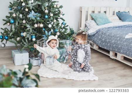 Two little sister girls open their gifts at the Christmas tree in the morning on the deck Two little sister girls open their gifts at the Christmas tree in the morning on the deck 122793383