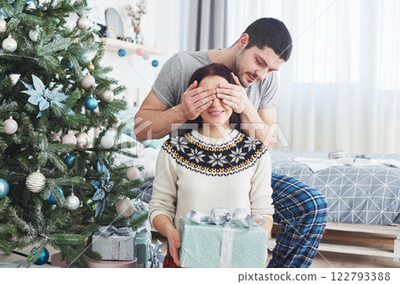 Young couple celebrating Christmas. A man suddenly presented a present to his wife. The concept of family happiness and well-being Young couple celebrating Christmas. A man suddenly presented a present to his wife. The concept of family happiness and well-being 122793388