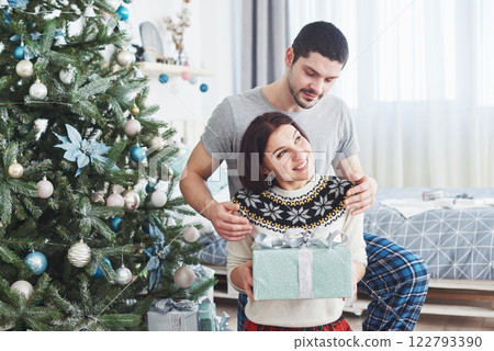Young couple celebrating Christmas. A man suddenly presented a present to his wife. The concept of family happiness and well-being Young couple celebrating Christmas. A man suddenly presented a present to his wife. The concept of family happiness and well-being 122793390