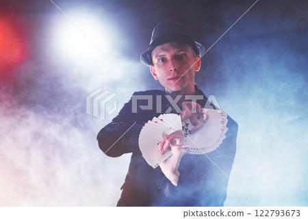 Magician showing trick with playing cards. Magic or dexterity, circus, gambling. Prestidigitator in dark room with fog Magician showing trick with playing cards. Magic or dexterity, circus, gambling. Prestidigitator in dark room with fog 122793673